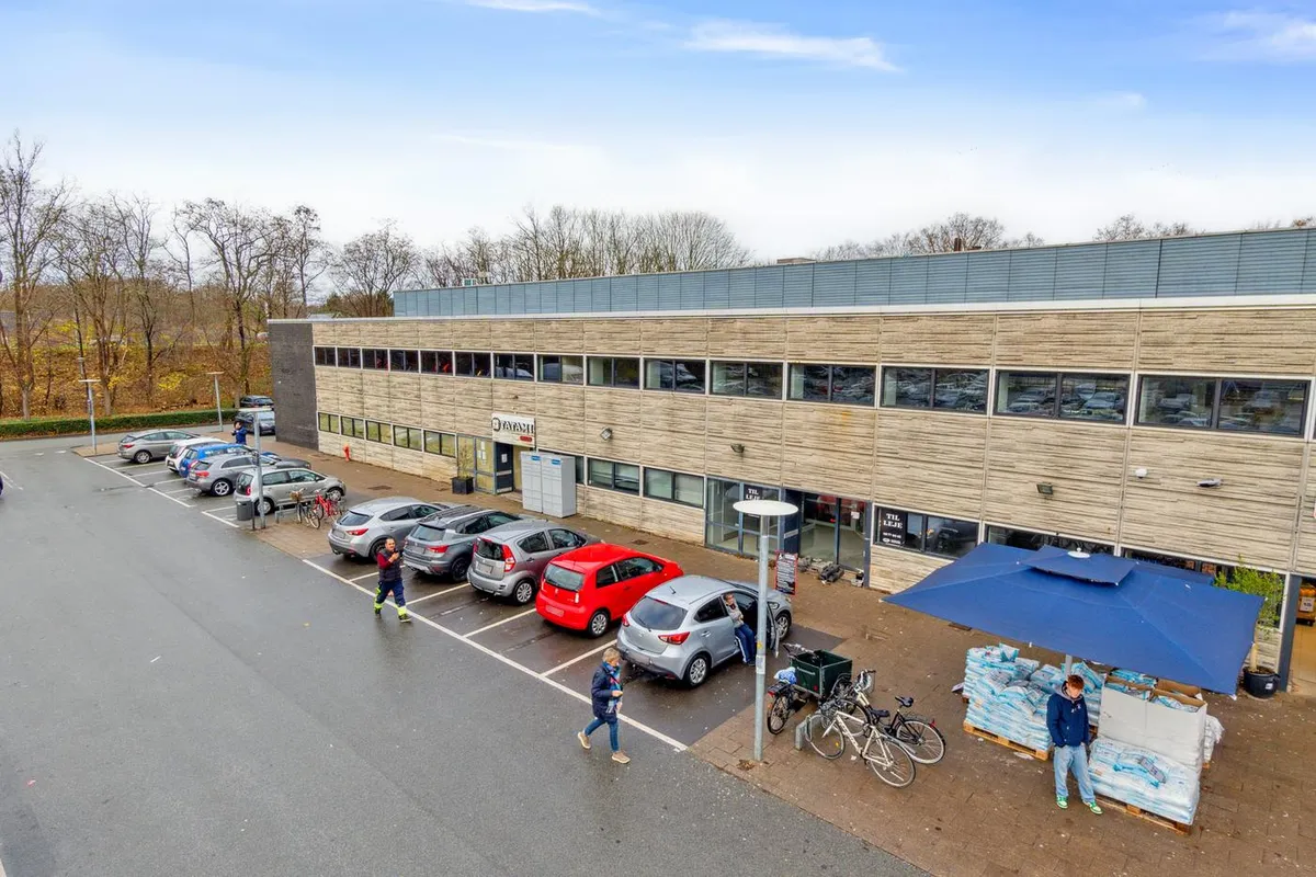 Parkerede biler foran et kontorbygning i Gyngemosen. Folk går forbi på gågaden, og der er en blå parasol med salgsvarer