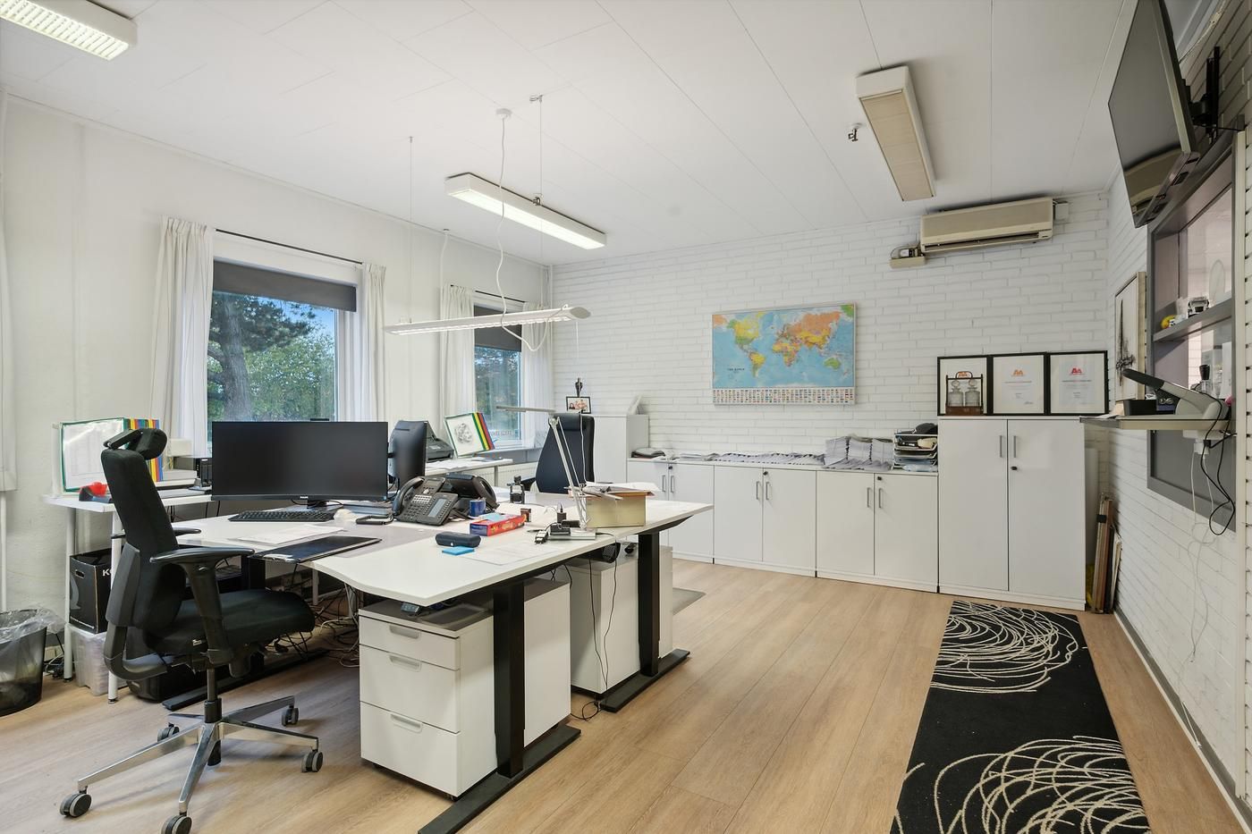 Kontorrum med skriveborde, computere, og kontormøbler, samt højt gulv og hvide vægge og lofter.