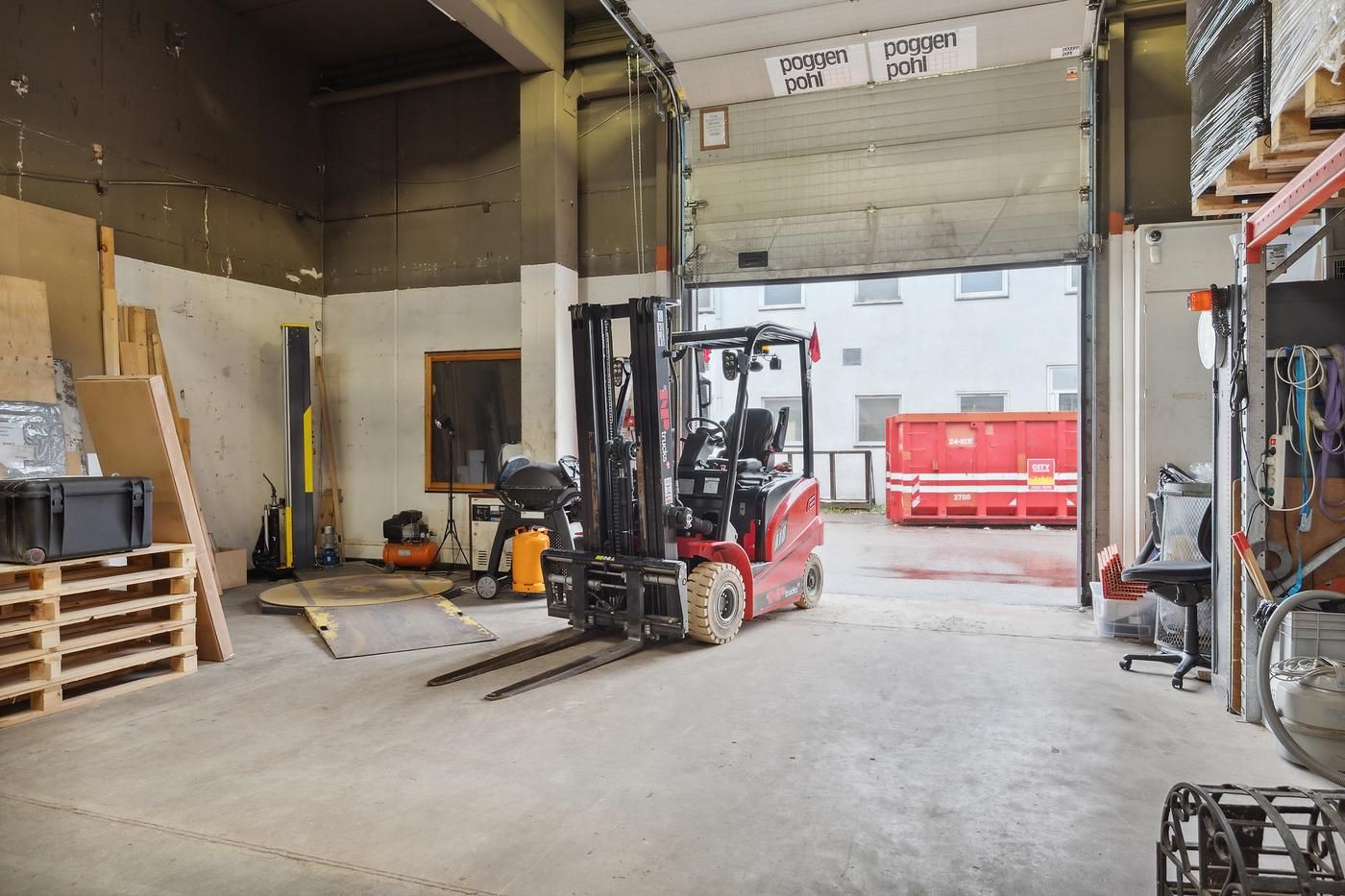 Et lagerrum med en forklæftningsmaskine, luftkompressor og træpaller, og en kørende gaffeltruck står ved åbningen mod en rød