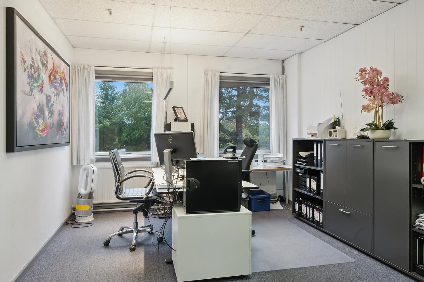 Kontor med computer, skrivebord, computer, stol, kabinet, blomster og maleri i et rum med vinduer og en hvid væg