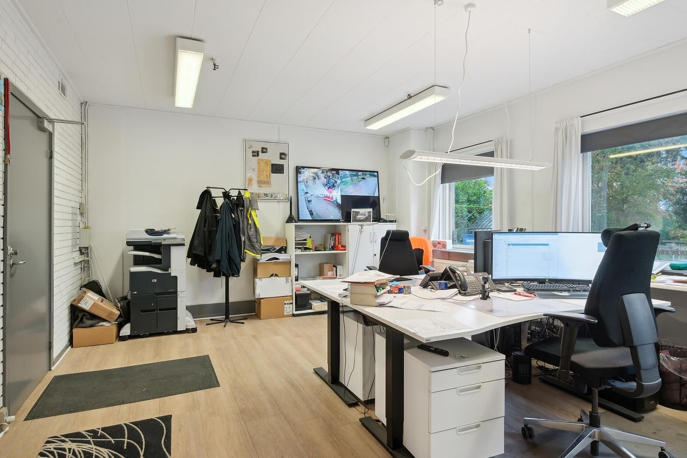 Kontor med højt gulv, hvide vægge, monitorer, skrivebord, skrivemaskine, køkkenbord, stole, og tøj på hængebolt