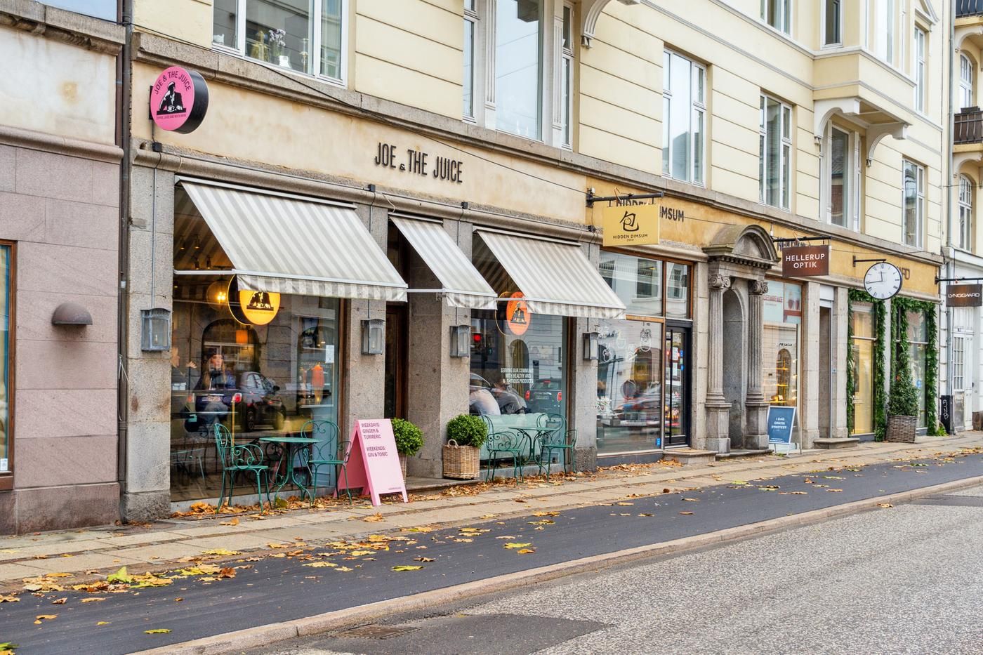 På en gågade med forretninger og røde blade er der en café med grønne stole og bord, samt en skrift på facaden med navnet
