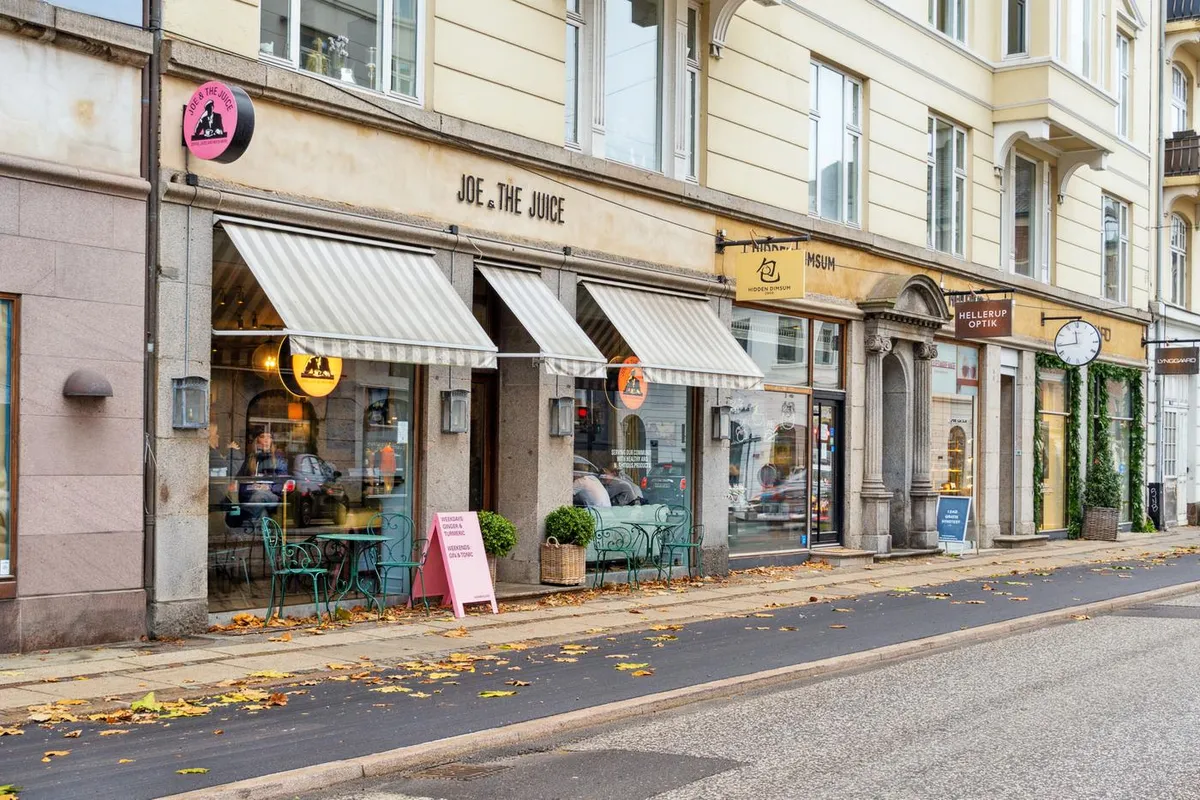 På en gågade med forretninger og røde blade er der en café med grønne stole og bord, samt en skrift på facaden med navnet