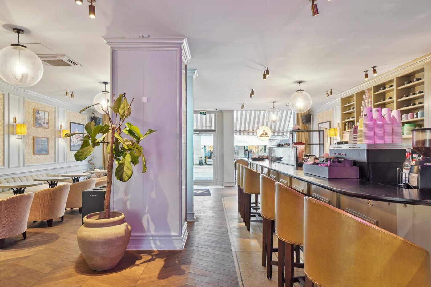 En stilfuld café med en stor plante, lyse stolpelampe, gulve og møbler i pastelfarver og en bar med kaffemaskine og