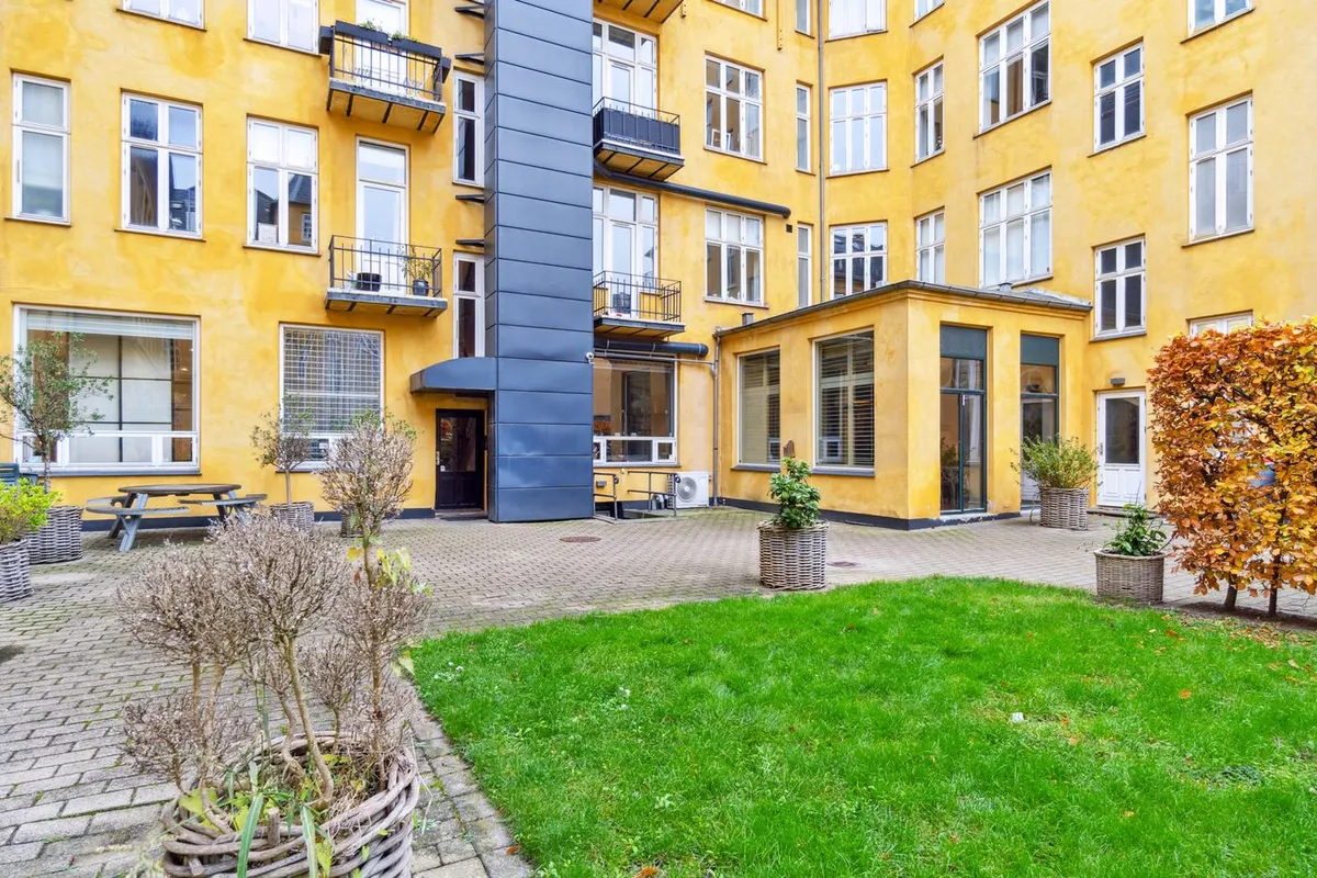 Forside til en pæn lejlighed med gul facade, to terrasser, gårdhave med græsplæne og træplanter samt en picnicbord og bænk