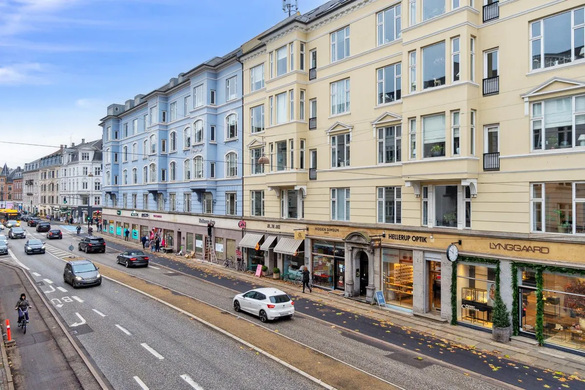 Strømforsyningsledninger over hovedgade med biler og cyklister samt bygninger med butikker og etableringer som Hellestrup