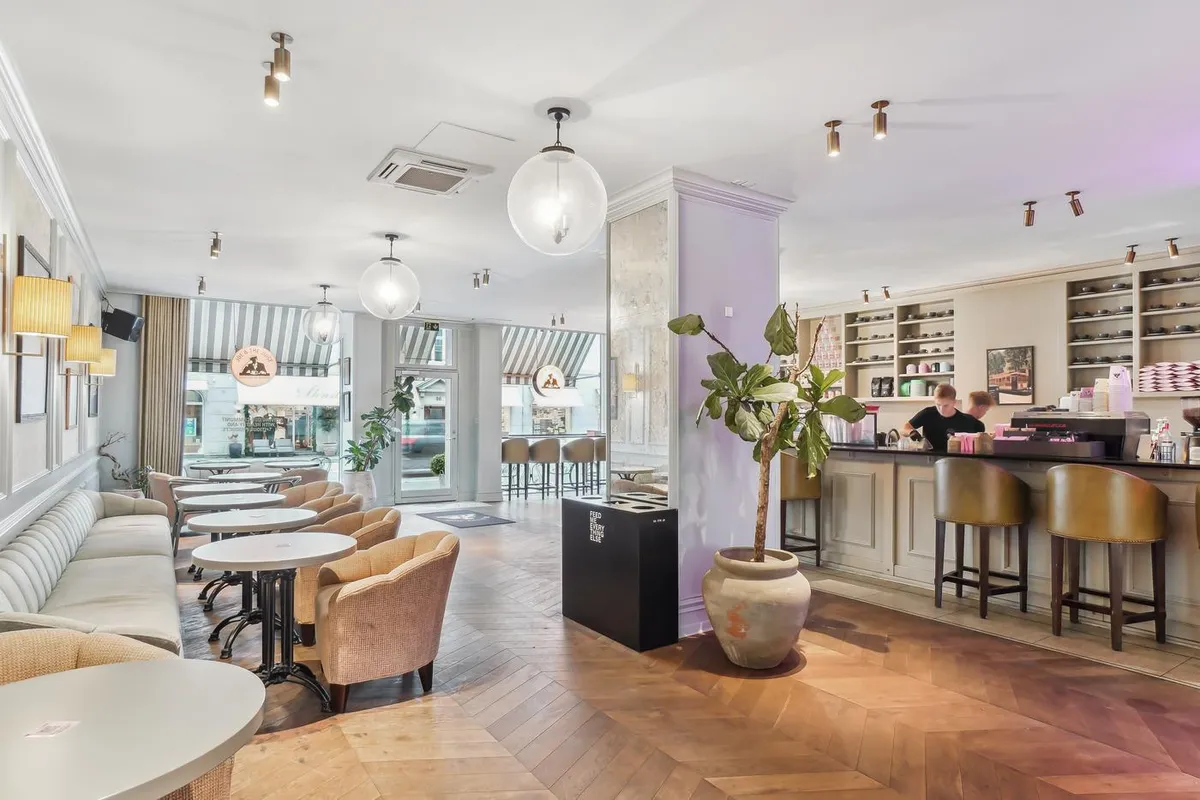 Inde i kafé med højt loft, hyggeligt møbelsæt og høj kvalitets trægulv. Bar og sofaer, samt lyse gulvlampe.