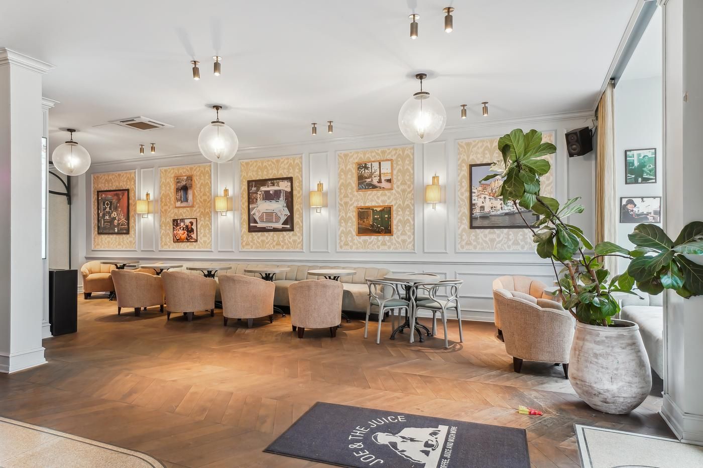 Møbelserie med bord og stole i café med højloft og plante i trægulv. Foto af café med loftslampe og malerier på væggen