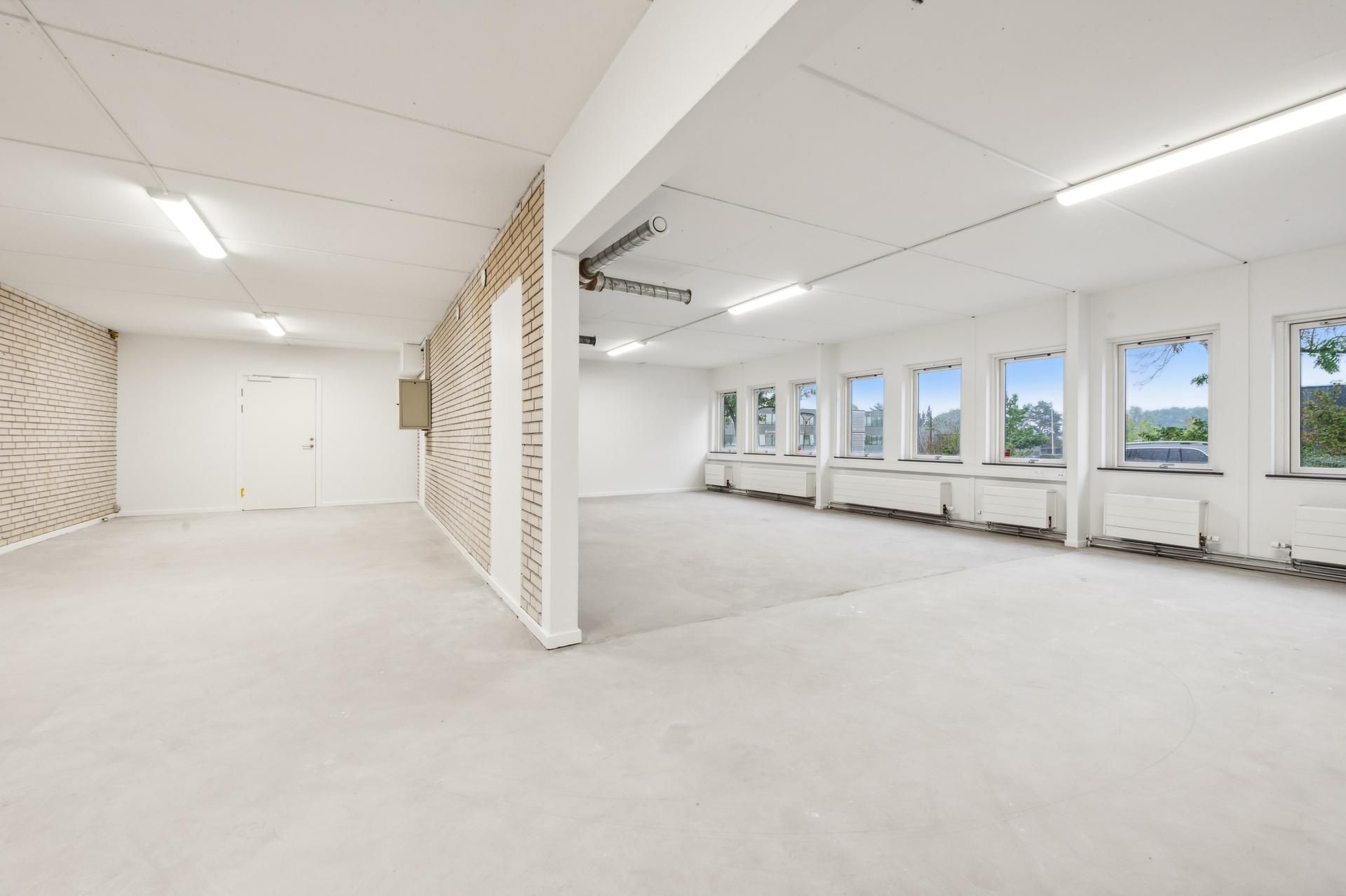 En stor lagerhal med hvide vægge, højt til loftet og mange vinduer med udsigt til skoven og bygninger i baggrunden