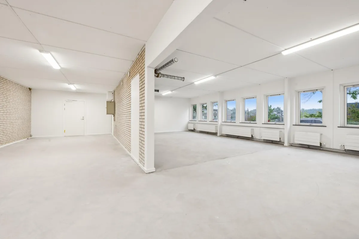 En stor lagerhal med hvide vægge, højt til loftet og mange vinduer med udsigt til skoven og bygninger i baggrunden