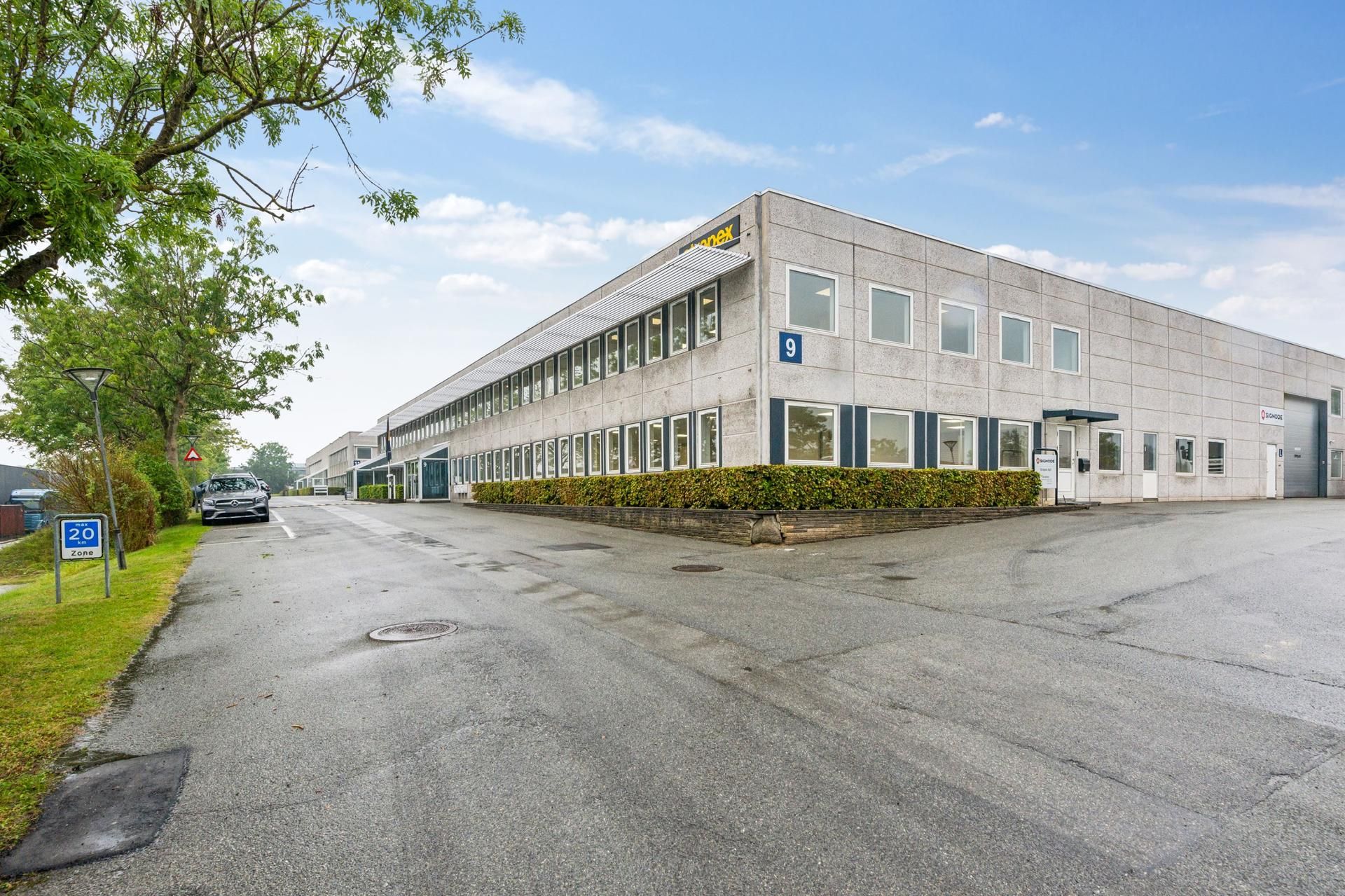 Lagerbygning med store vinduer, parkeringsplads og en bilparkeret Mercedes foran. En 20 km/t skilt og en stor trægruppe