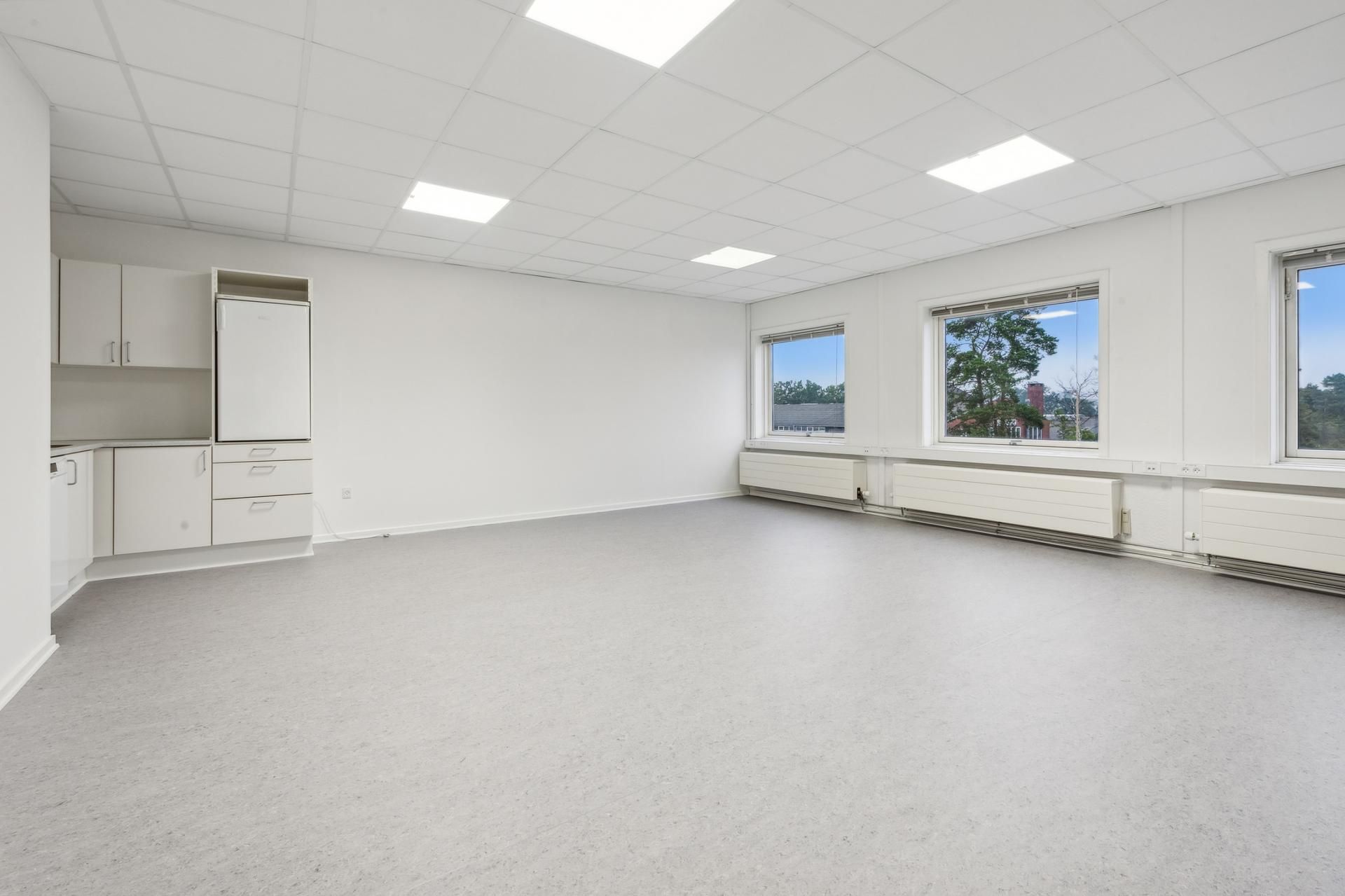 Hvidt lokale med højt til loftet, hvide vægge og højt gulv med lysepunkter og tre store vinduer med udsigt til træer