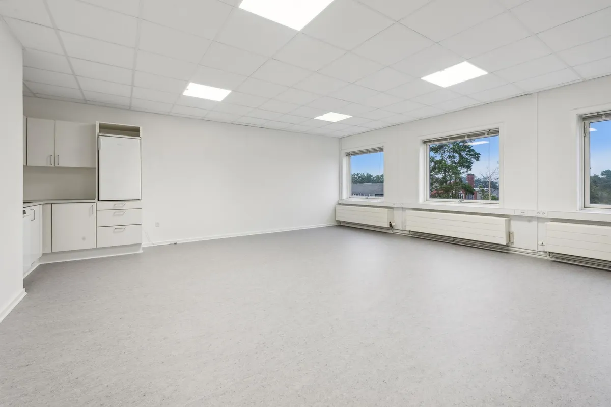 Hvidt lokale med højt til loftet, hvide vægge og højt gulv med lysepunkter og tre store vinduer med udsigt til træer