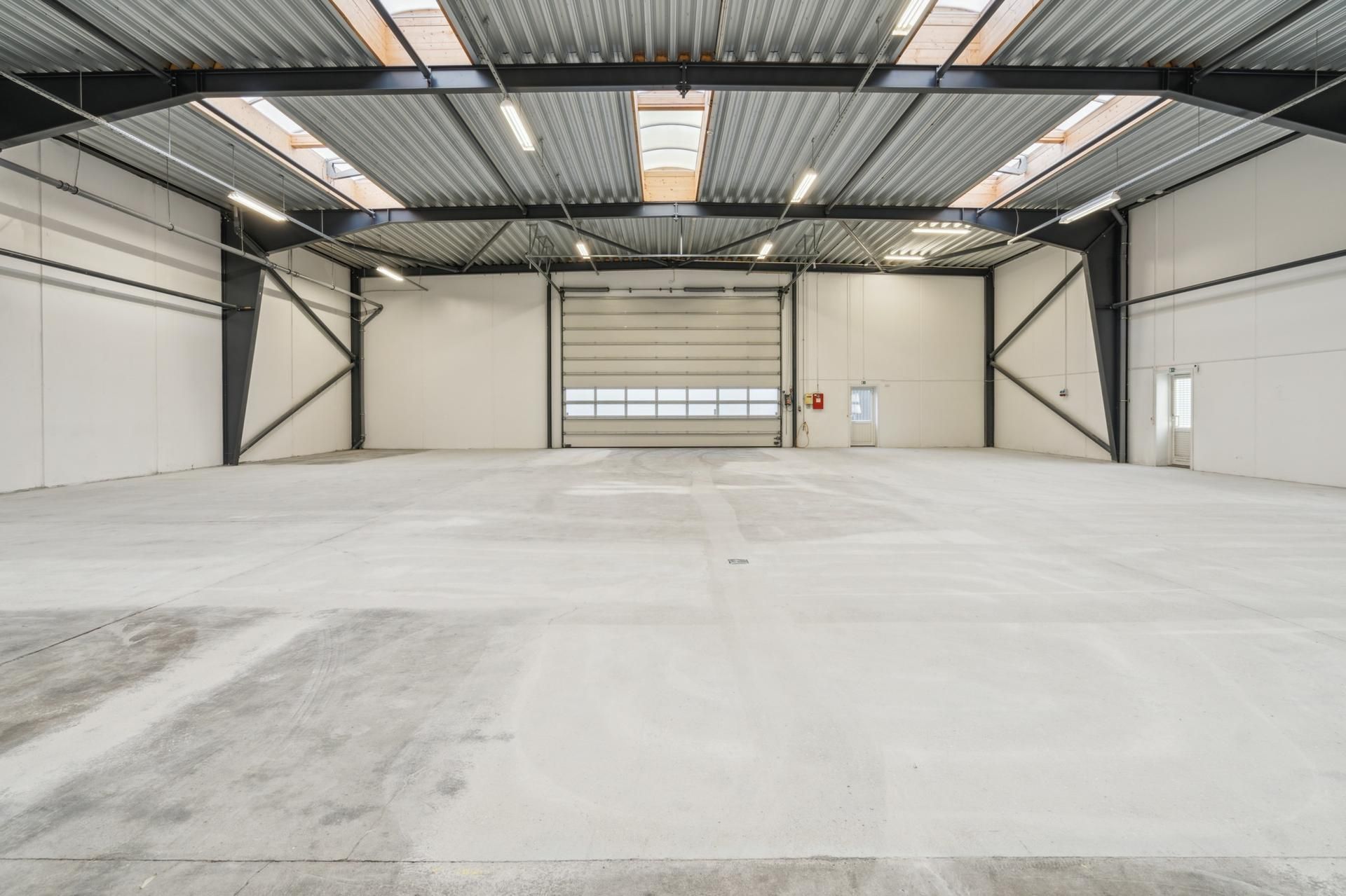 En stor, tom hal med lyse loftsvinduer og hvide vægge. Værkstedet har store glasdøre, lysekroner og et rødt brandslukker.