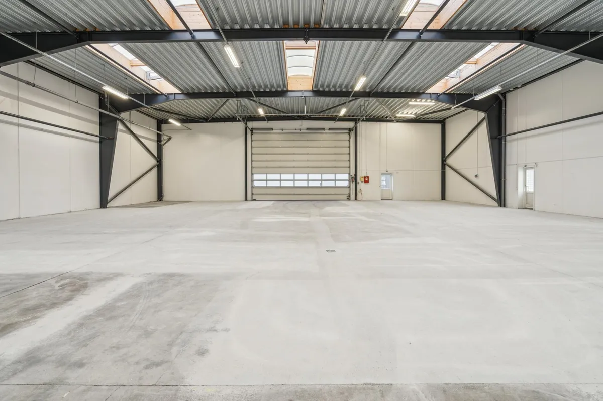 En stor, tom hal med lyse loftsvinduer og hvide vægge. Værkstedet har store glasdøre, lysekroner og et rødt brandslukker.