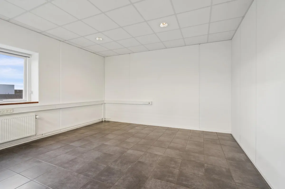Ledig kontorplads med hvidt loft, hvide vægge og en stor, tom væg med vindue. Gulvet er dækket med grå fliser.