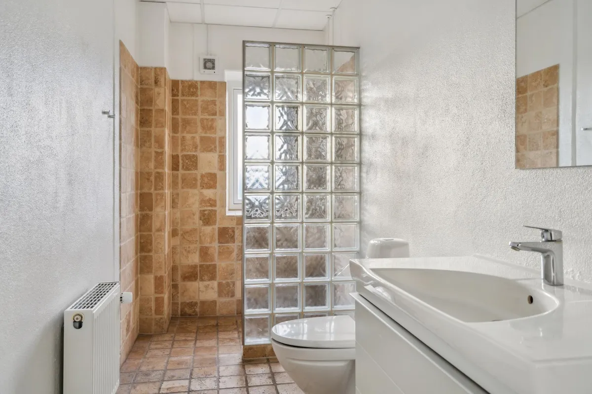 Lejlighedens badeværelse med brusekabine, hvidt toilet, lysebrune og lyse beige fliser og en hvid radiator