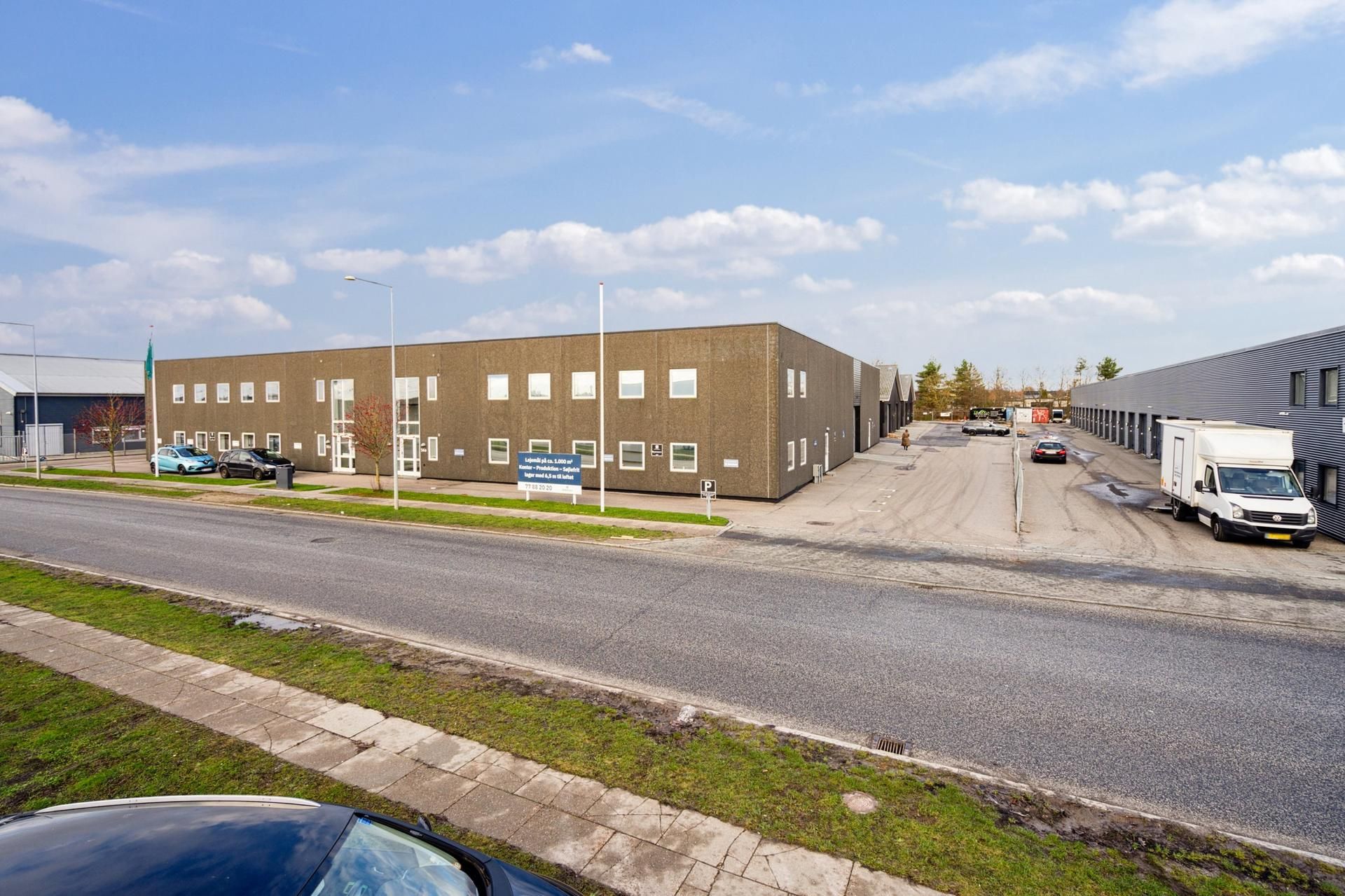 Forretningslokaler med parkeringsplads, hvor en person går og to biler er parkeret, samt en hvid lastvogn parkeret i