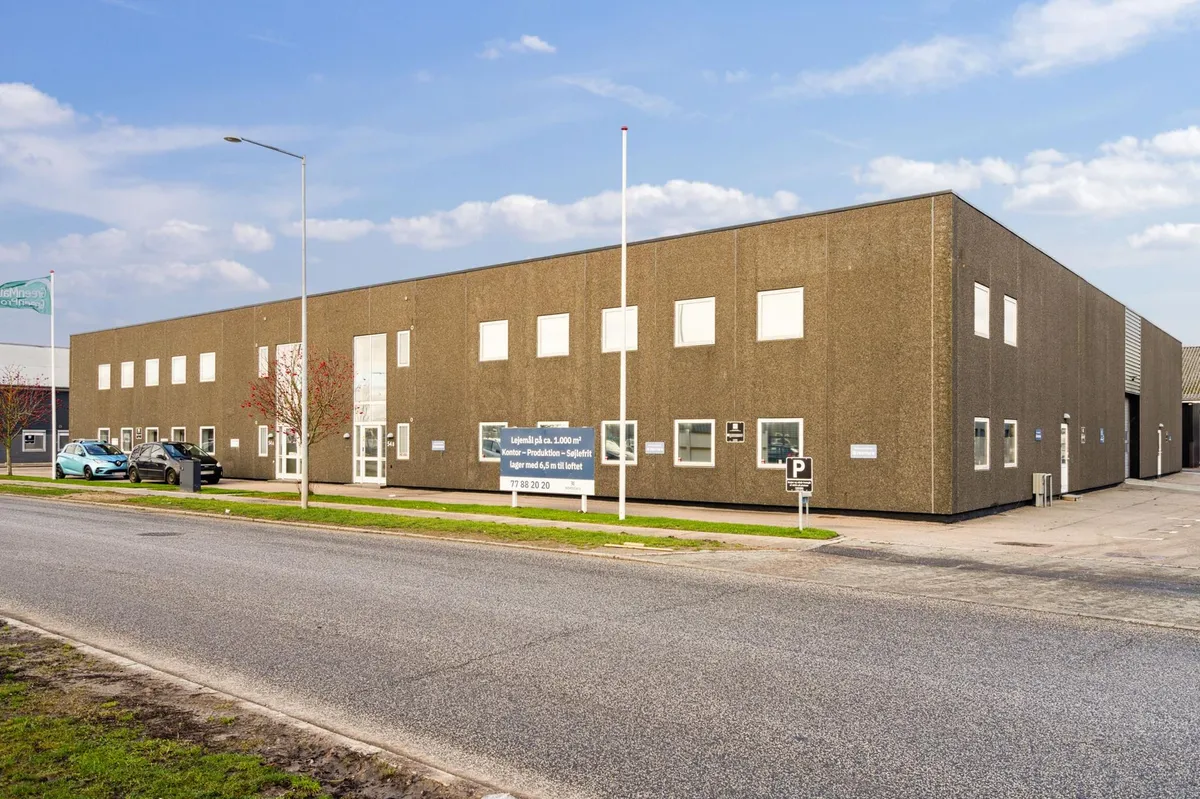 Et stort, brunlagt bygning med mange vinduer og skilte med tekst. Parkerede biler og grønne buske foran. En parkeringsskilt