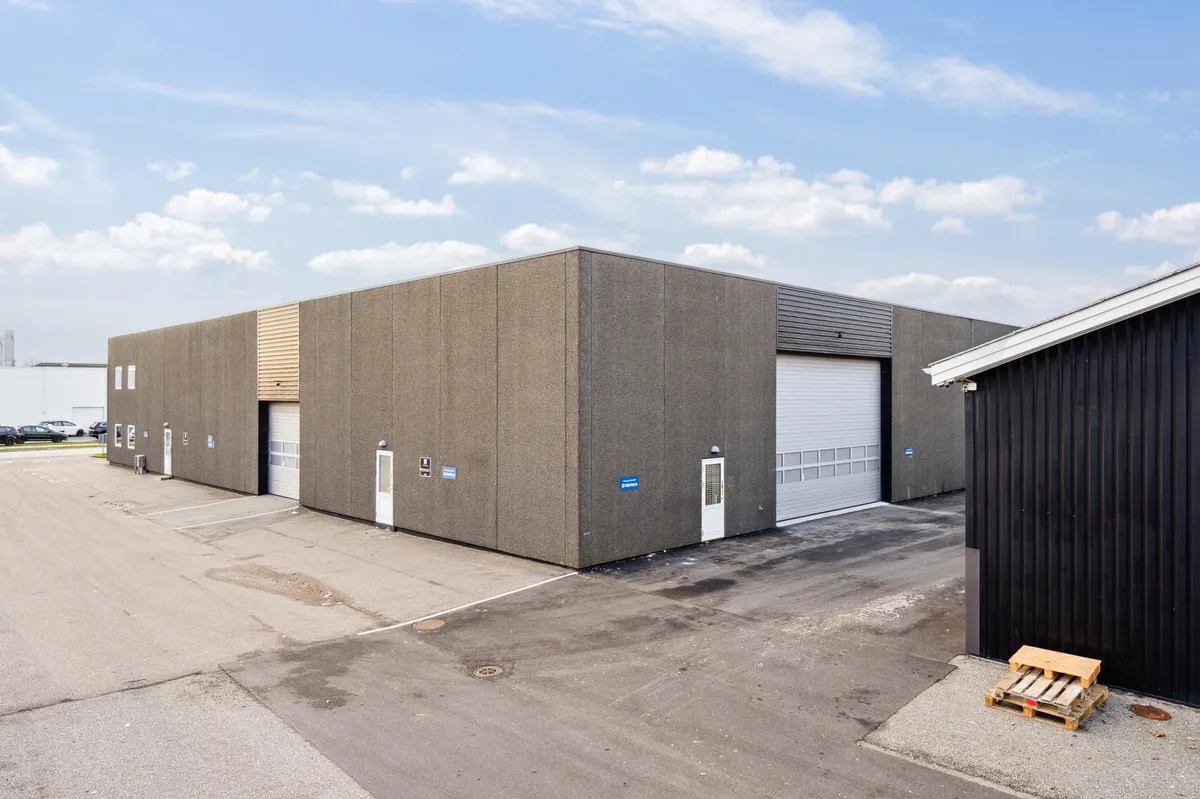 To industribygninger med hvide døre og garageporte og en stor parkeringsplads foran