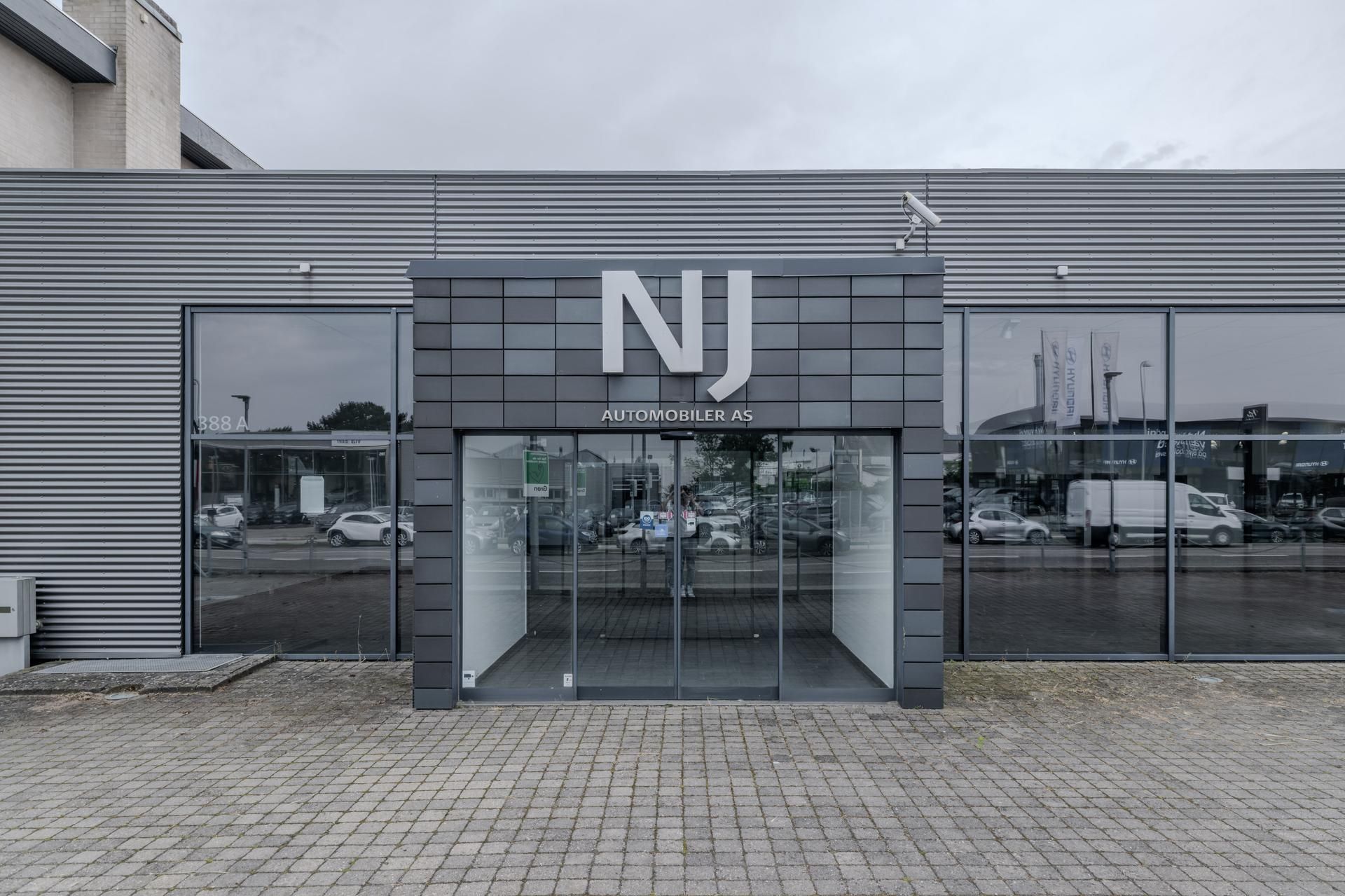 Indgang til NJ Automobiler AS med store glasdøre og refleksioner af biler, bygninger og mennesker