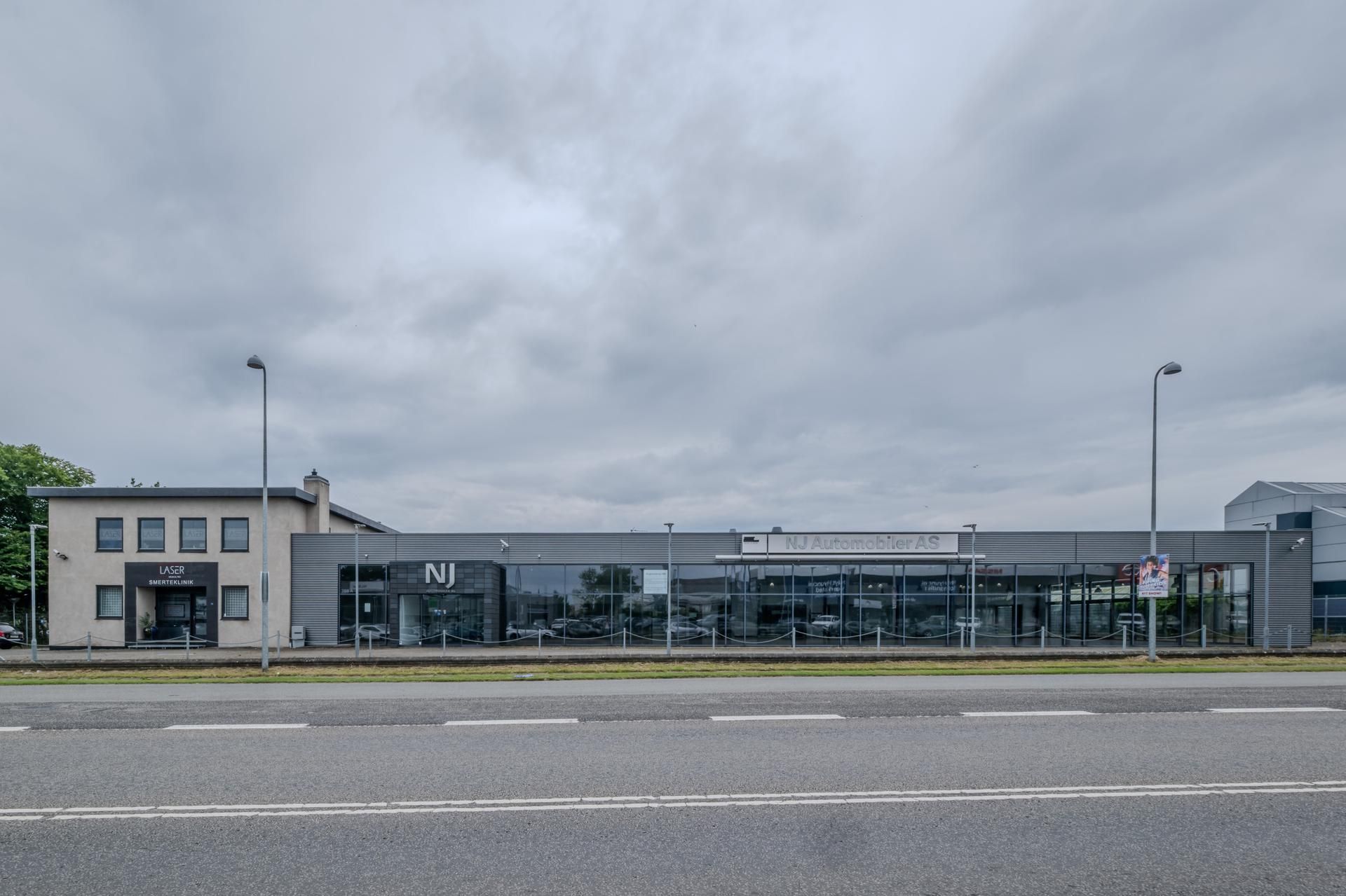 Bygning med glasfront og skilte, der læser NJ Automobiler AS. To søjler med lamper, større bygning til højre, små bygninger