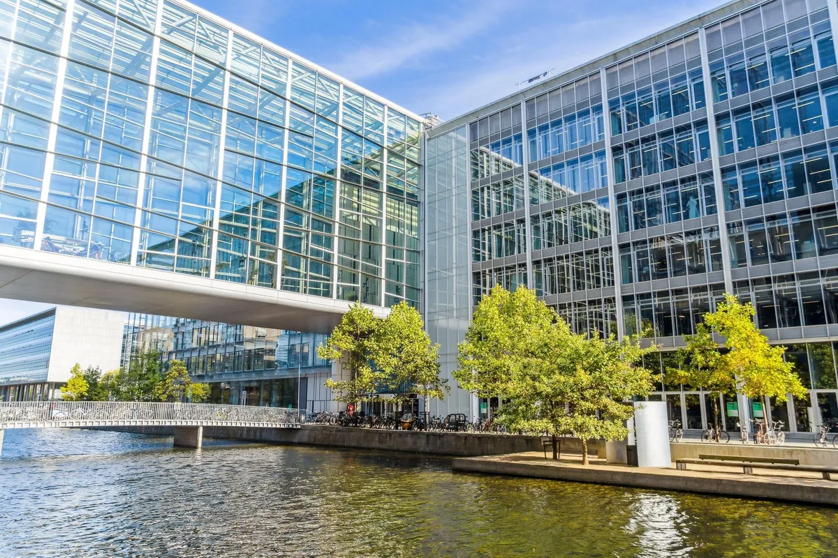 Moderne bygning med glasfasade ved en kanal og en bro foran bygningen med træer langs siderne