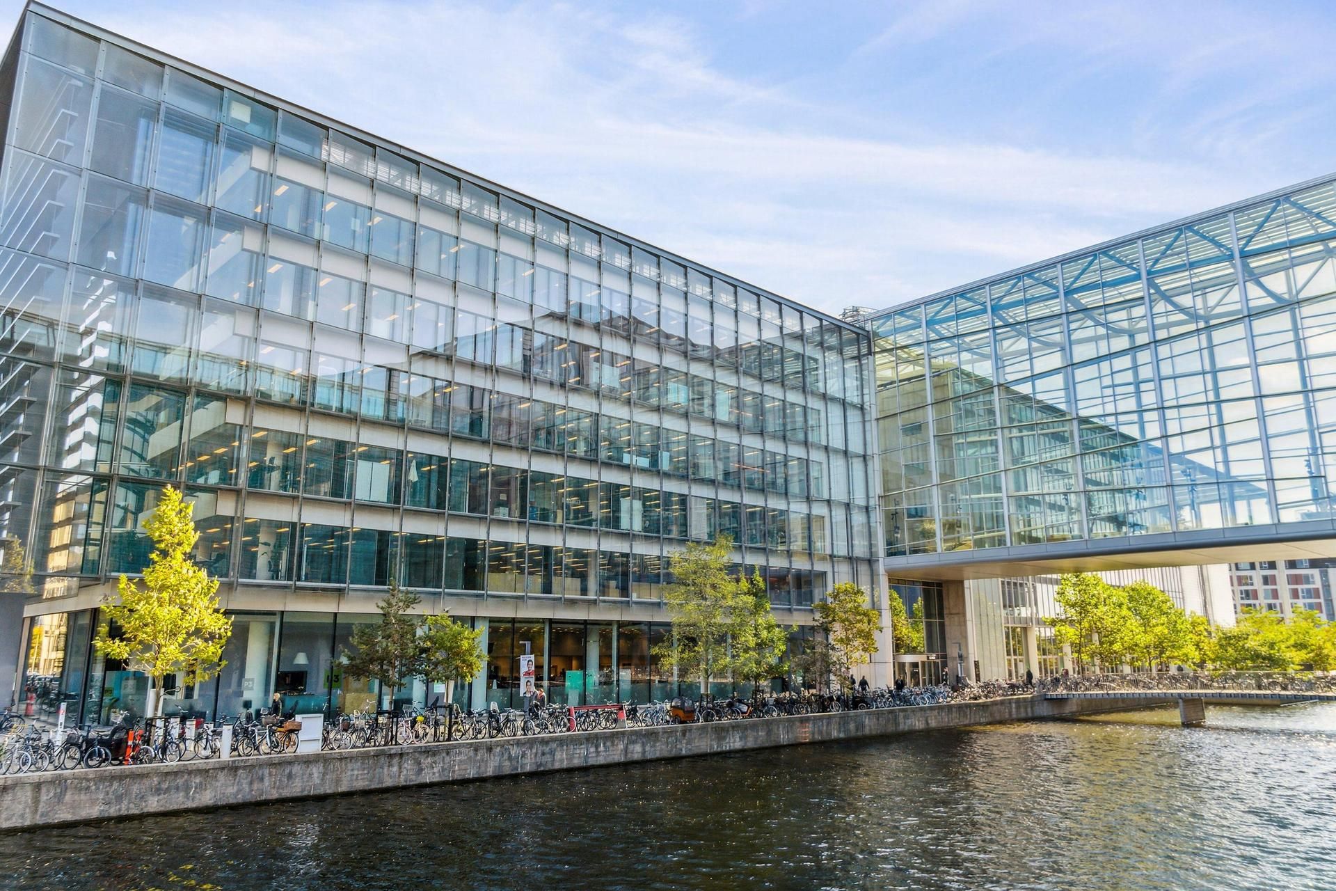 Moderne bygning med glasfassade og slanke søjler, parkerede cykler, fredelig vandoverflade, grønne træer og et brobyggeri