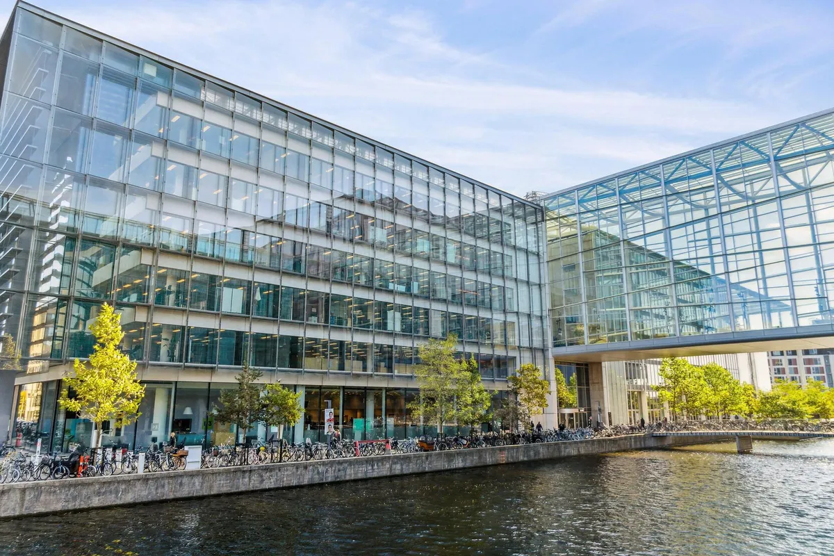 Moderne bygning med glasfassade og slanke søjler, parkerede cykler, fredelig vandoverflade, grønne træer og et brobyggeri