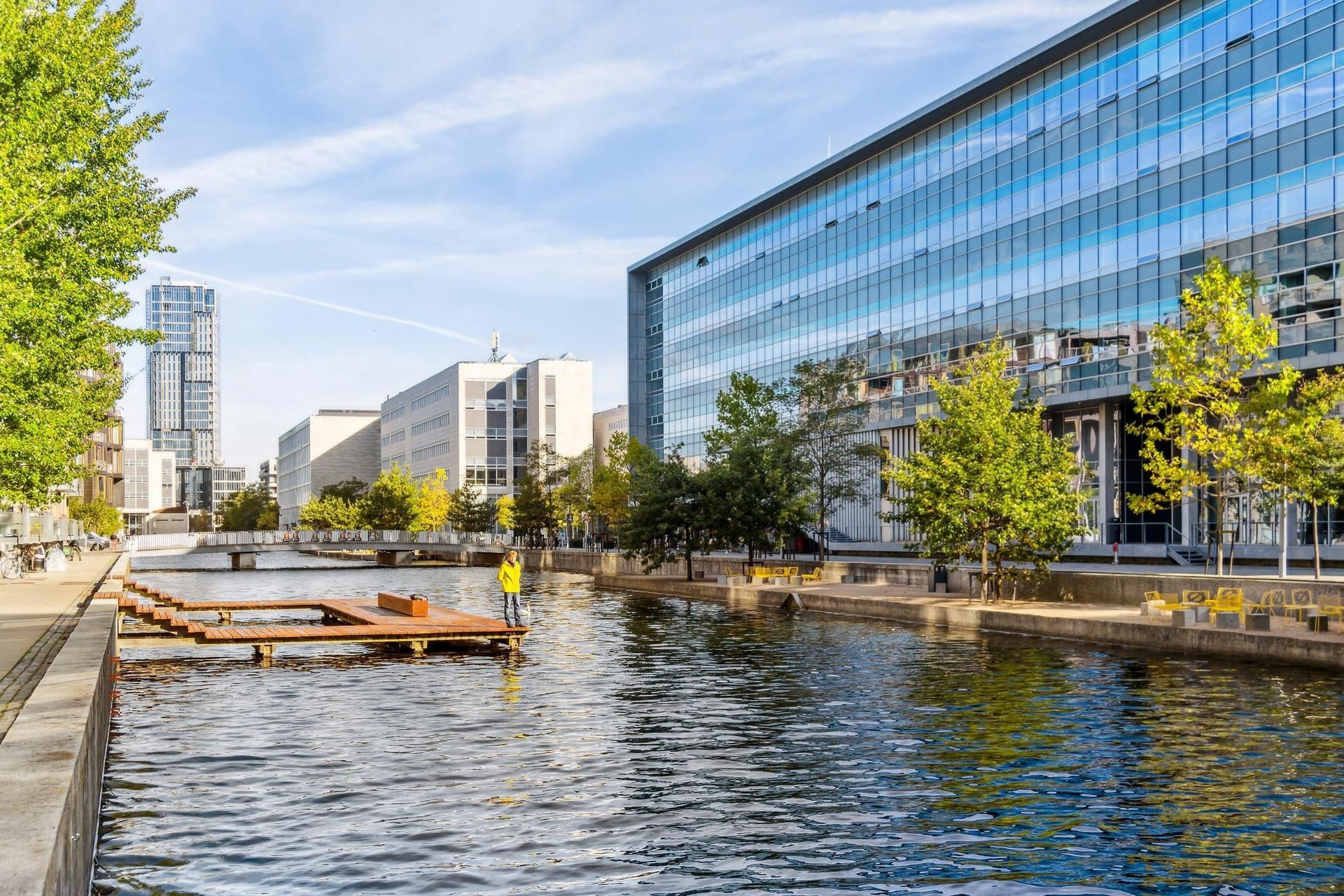 En bygning med glasvinduer står på en bro, der går over en vandvej, omgivet af træer og grønne planter