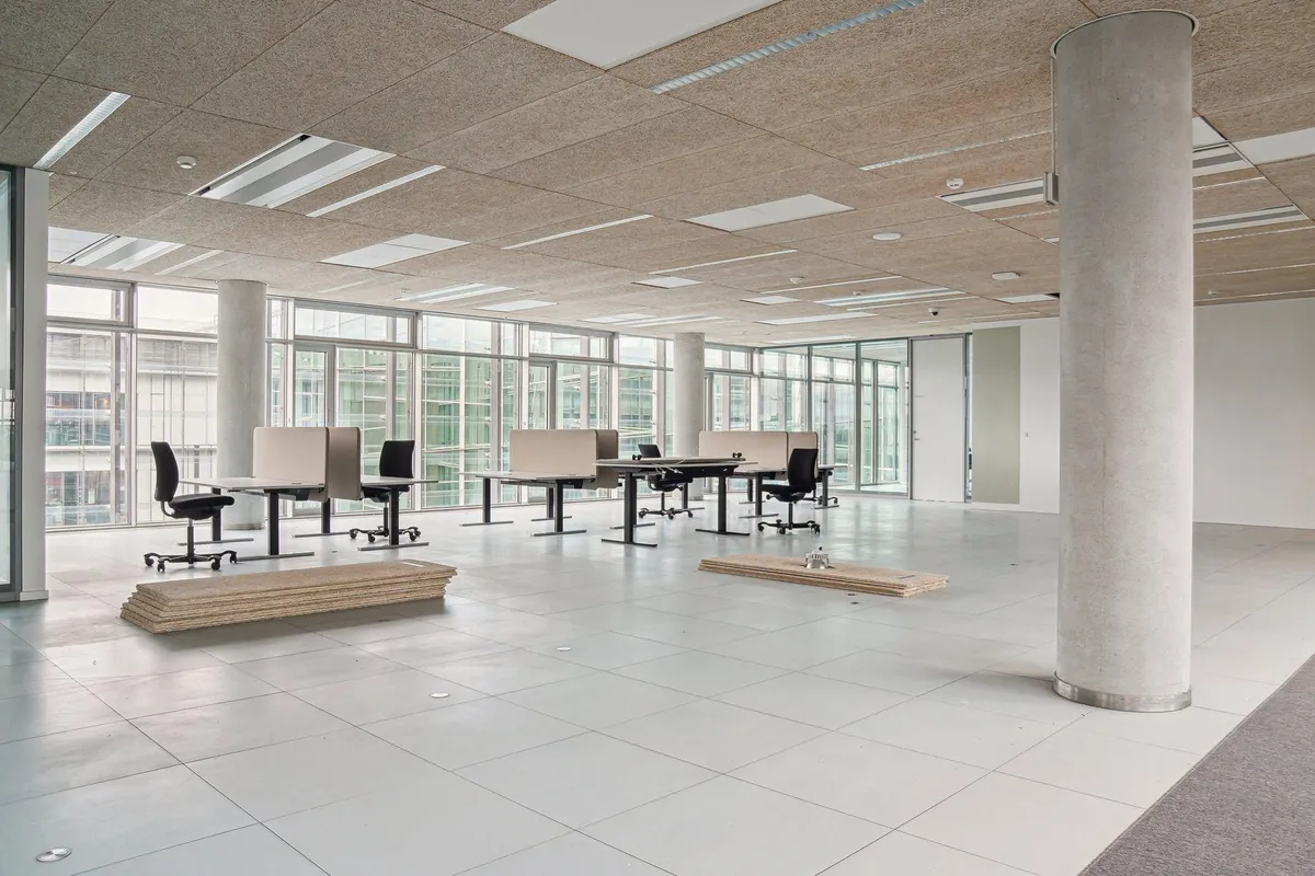 Moderne kontorrum med hvide fliser, glasvinduer, høje lofter og søjler, flere borde og stole samt skarpe linjer og lyse