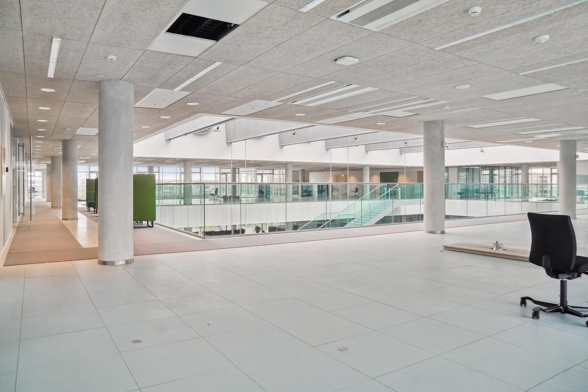 Moderne bygning med højloftede rum, hvid gulvbelægning, glasvinduer, og kontormøbler i hjørner.