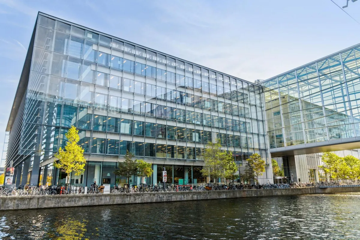 Mange cykler parkeret ved en stor bygning med glasvinduer og udsigt over en vandkanal med fredelig vandoverflade