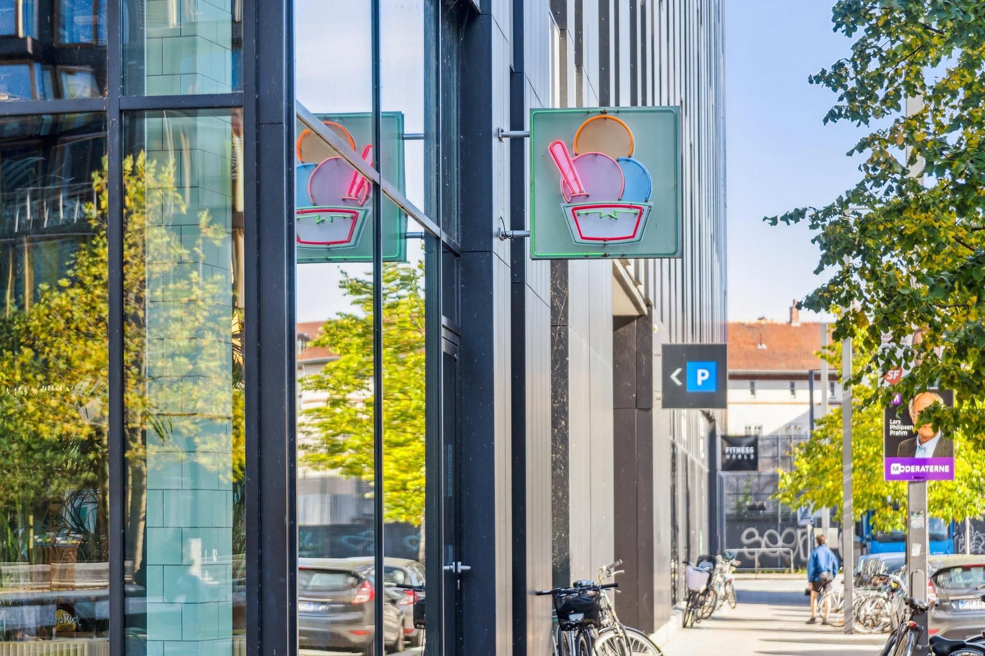 DR-byen i København med neonskilte og slikkopper, cykler, mennesker og biler parkeret langs gaden med busstop og annoncer