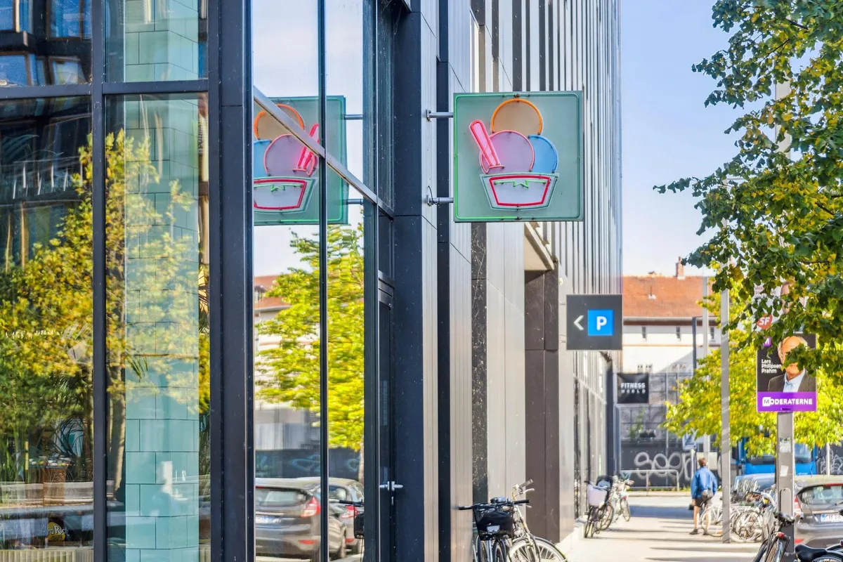 DR-byen i København med neonskilte og slikkopper, cykler, mennesker og biler parkeret langs gaden med busstop og annoncer