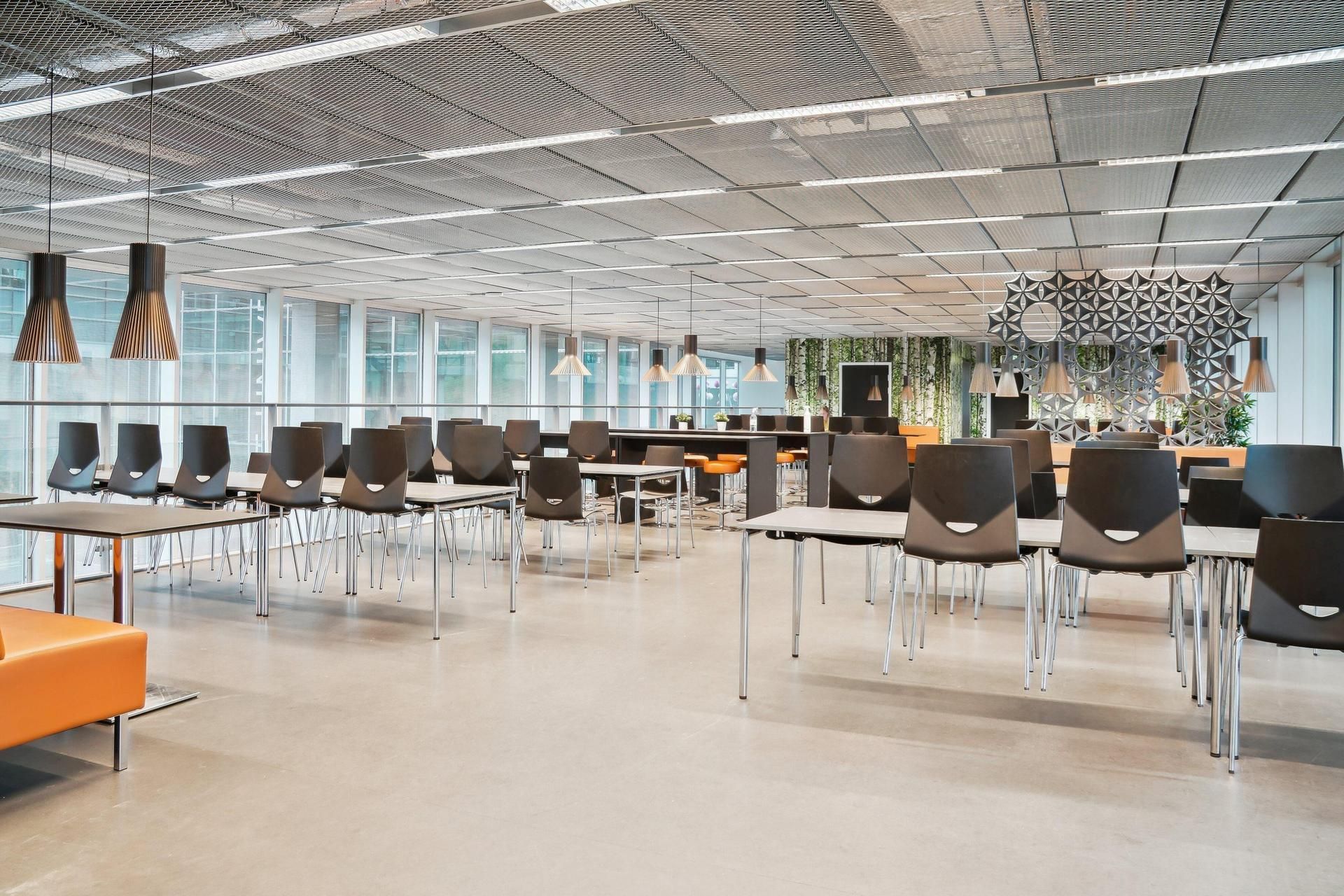 Værelse med store vinduer, lyse lofter og lysekroner, borde og stole, orange sofa og væg med en dekoreret panel