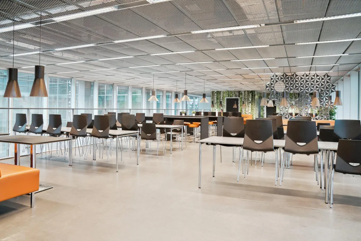 Værelse med store vinduer, lyse lofter og lysekroner, borde og stole, orange sofa og væg med en dekoreret panel