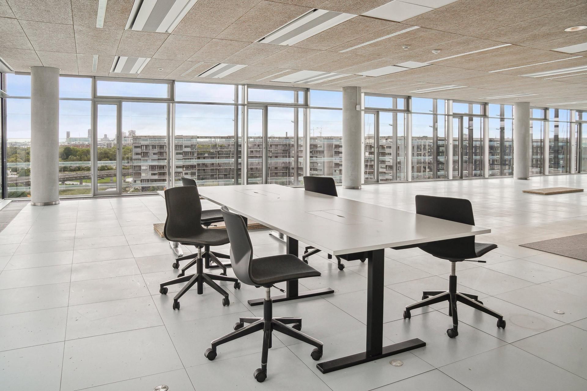 Moderne mødelokale med høje glasvinduer og udsigt over byen. Lang hvid bord med fire stole. Ingen mennesker i rummet.