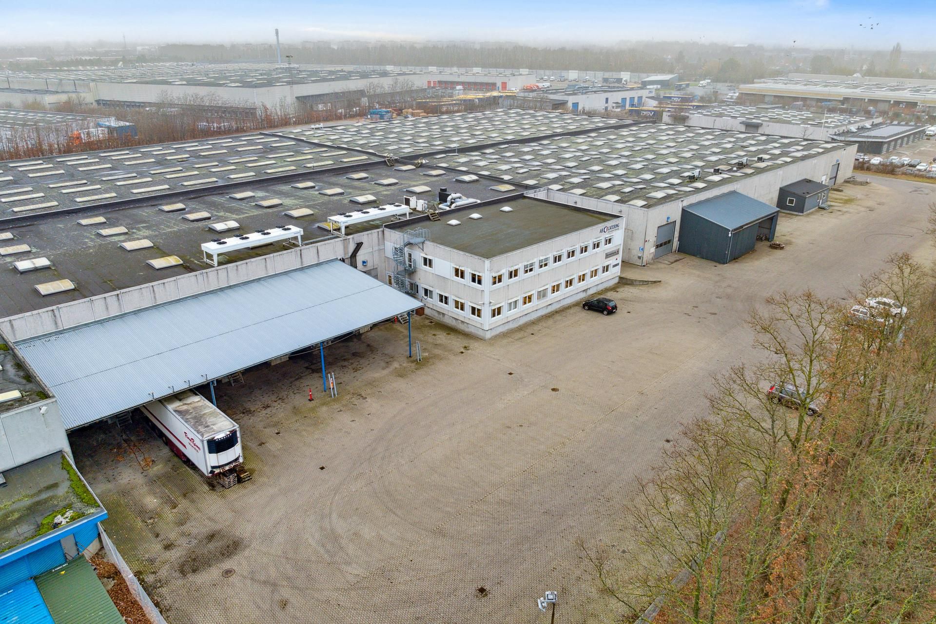 Fabriksanlæg med stort lager og mange vinduede bygninger samt biler og lastbiler parkeret i nærheden