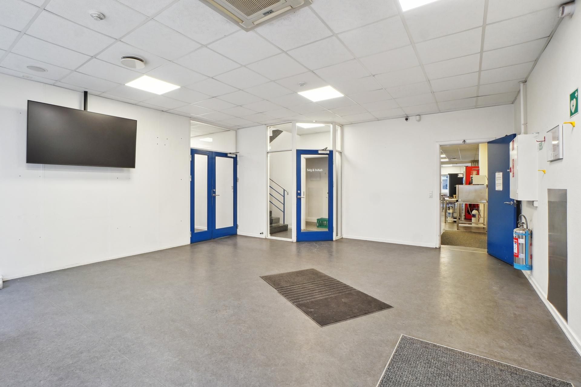 Industrialt rum med hvide vægge og gulve, to blå døre, et fjernsyn, luftkanaler og en ildslukker