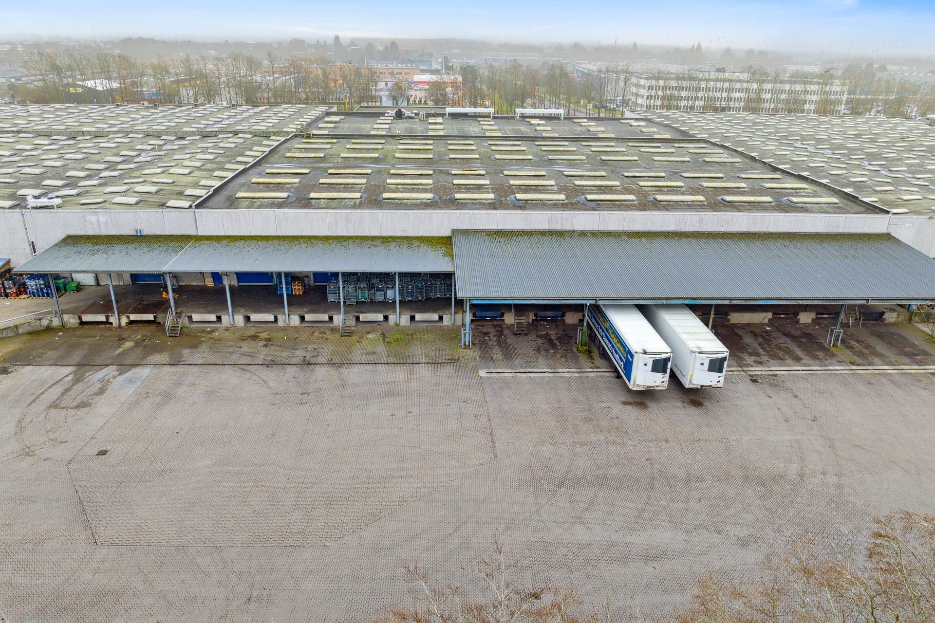 Fabrik med store lagerbygninger og lastbiler parkeret i rækker på asfaltvej, tæt på bygningskanten