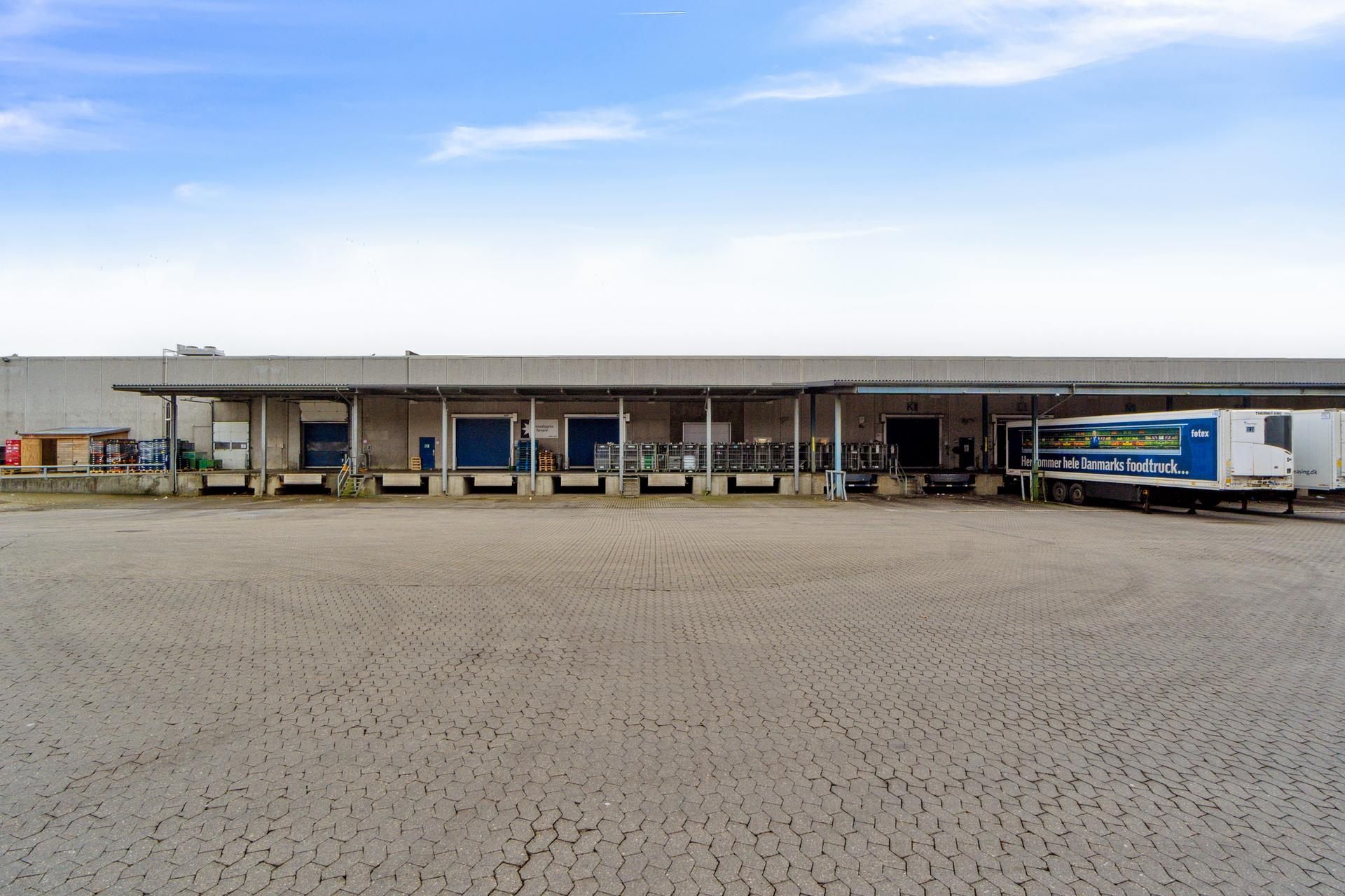 Lagerbygning med lastbilparkering og flere lagerporte i rødt og blåt, ligesom der er en hvid lastbilparkeret ved bygningen