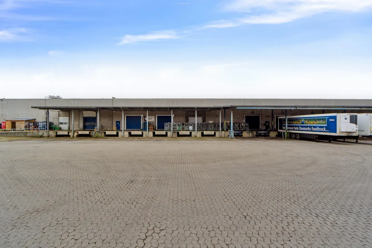 Lagerbygning med lastbilparkering og flere lagerporte i rødt og blåt, ligesom der er en hvid lastbilparkeret ved bygningen