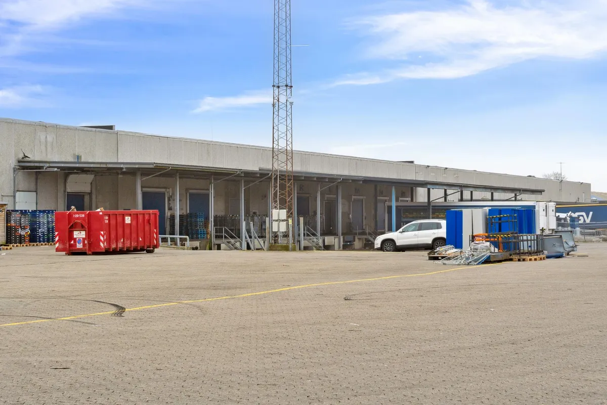 En stor industribygning med flere vinduer og røde containere parkeret foran og et parkerede køretøj