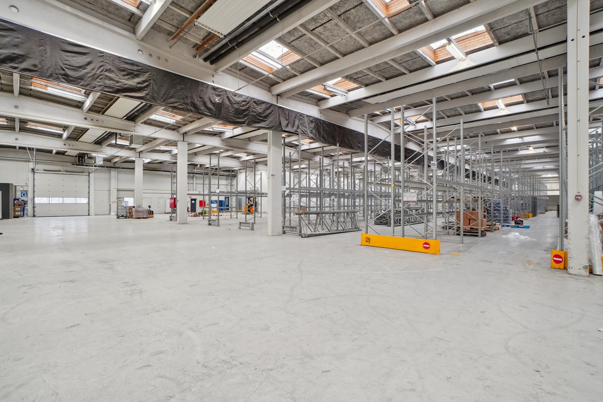 Et stort industrilager med metalliske opbevaringsstel og lyse lofter, fuld af paller og forskellige varer til fragt og lager.