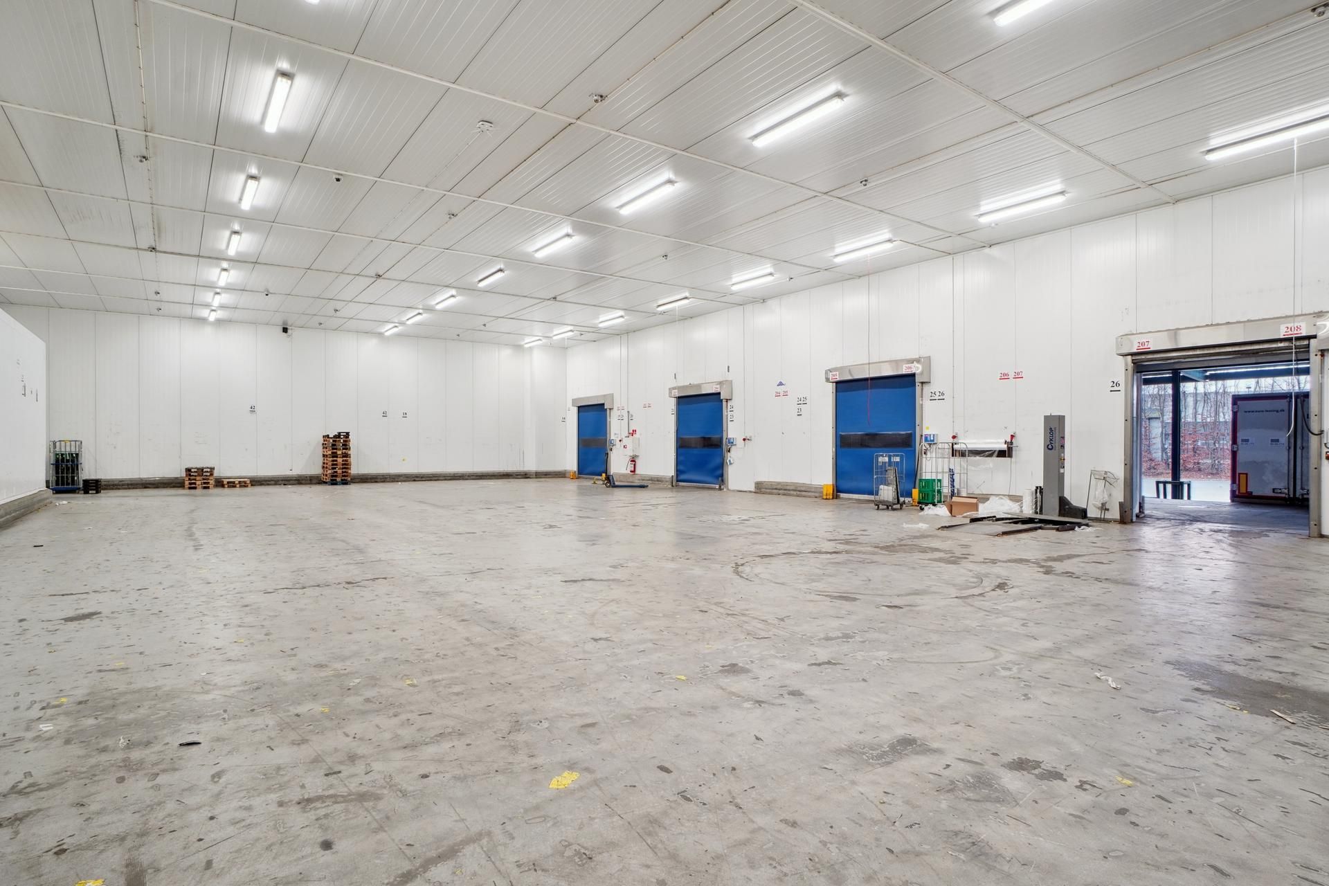 Lager med flere døre og to store åbninger, der fører ud til vejen. Der er stel og paller placeret i rummet, og der er flere