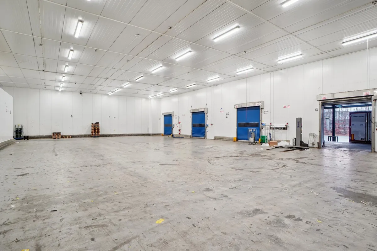 Lager med flere døre og to store åbninger, der fører ud til vejen. Der er stel og paller placeret i rummet, og der er flere