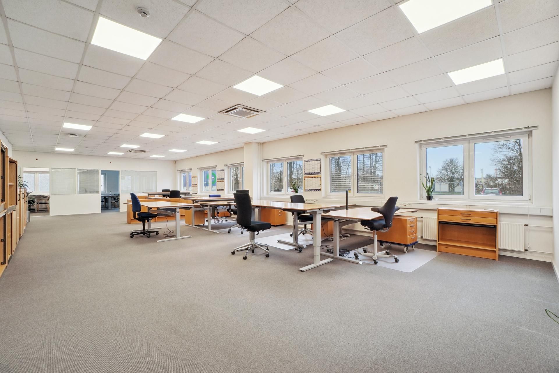 En tom kontorbygning med lyse gulve, lyse lofter og vinduer med gardiner, hvor der er flere bord og stol sæt samt en plante
