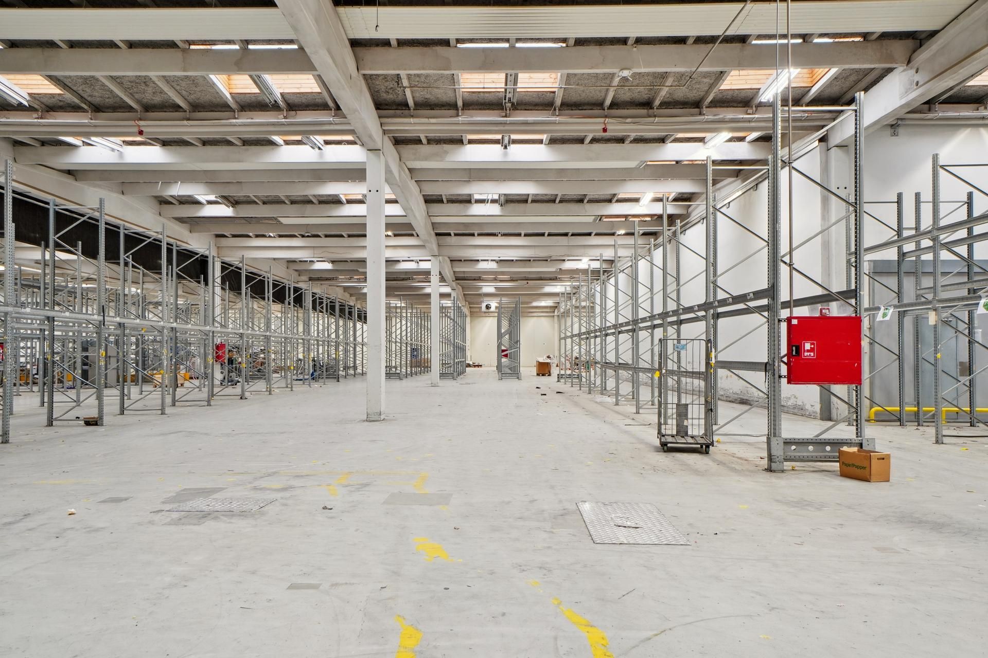 En stor, tom lagerhal med metalreoler og gule linjer på gulvet, der angiver adgangsveje og områder