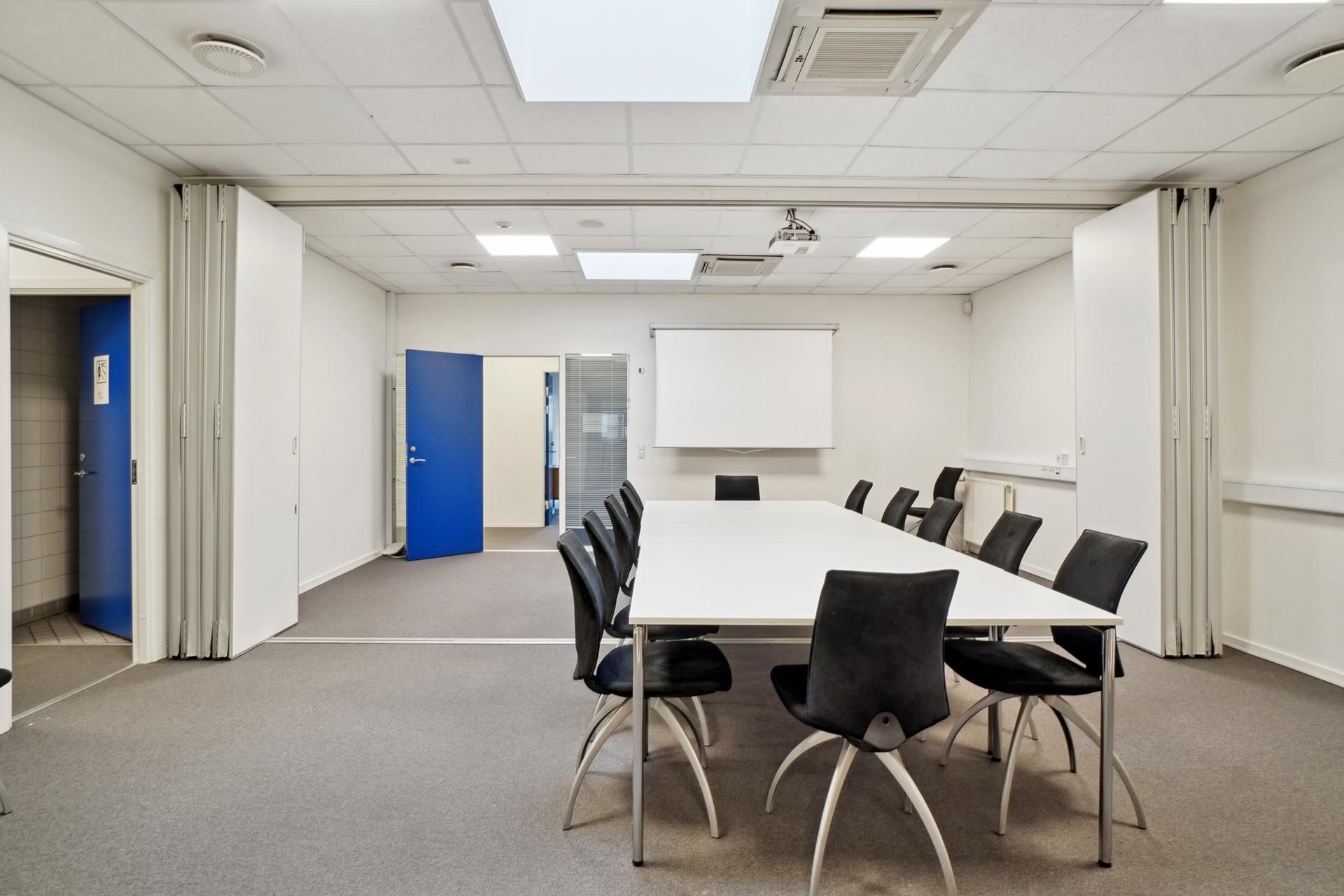 Langt konferencebord med flere stole omkring, hvidt bord og mørke stole, lyst hvidt loft, hvidt vægmaleri og projektor foran